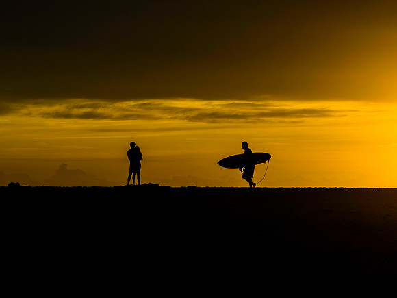 Sunrise silhouettes - lovers and a lone surfer heading out